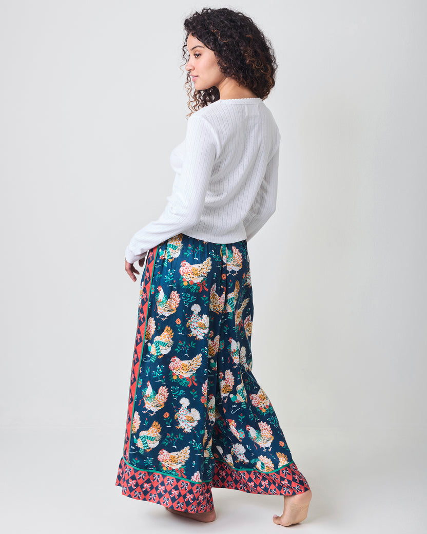 Back view of a person wearing the full-length Holiday Hens satin pants with a dark blue chicken print and red patterned borders.