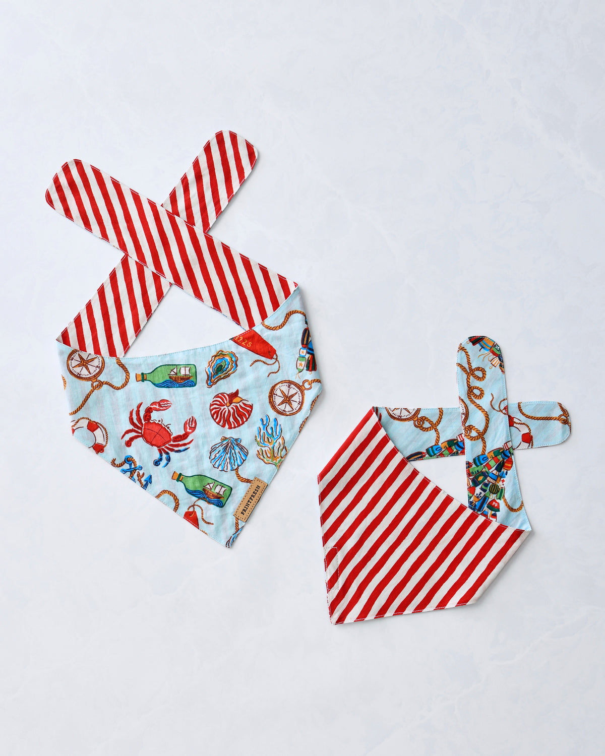 Two pet bandanas laid flat, each combining a pale blue nautical print with red-and-white striped ties.