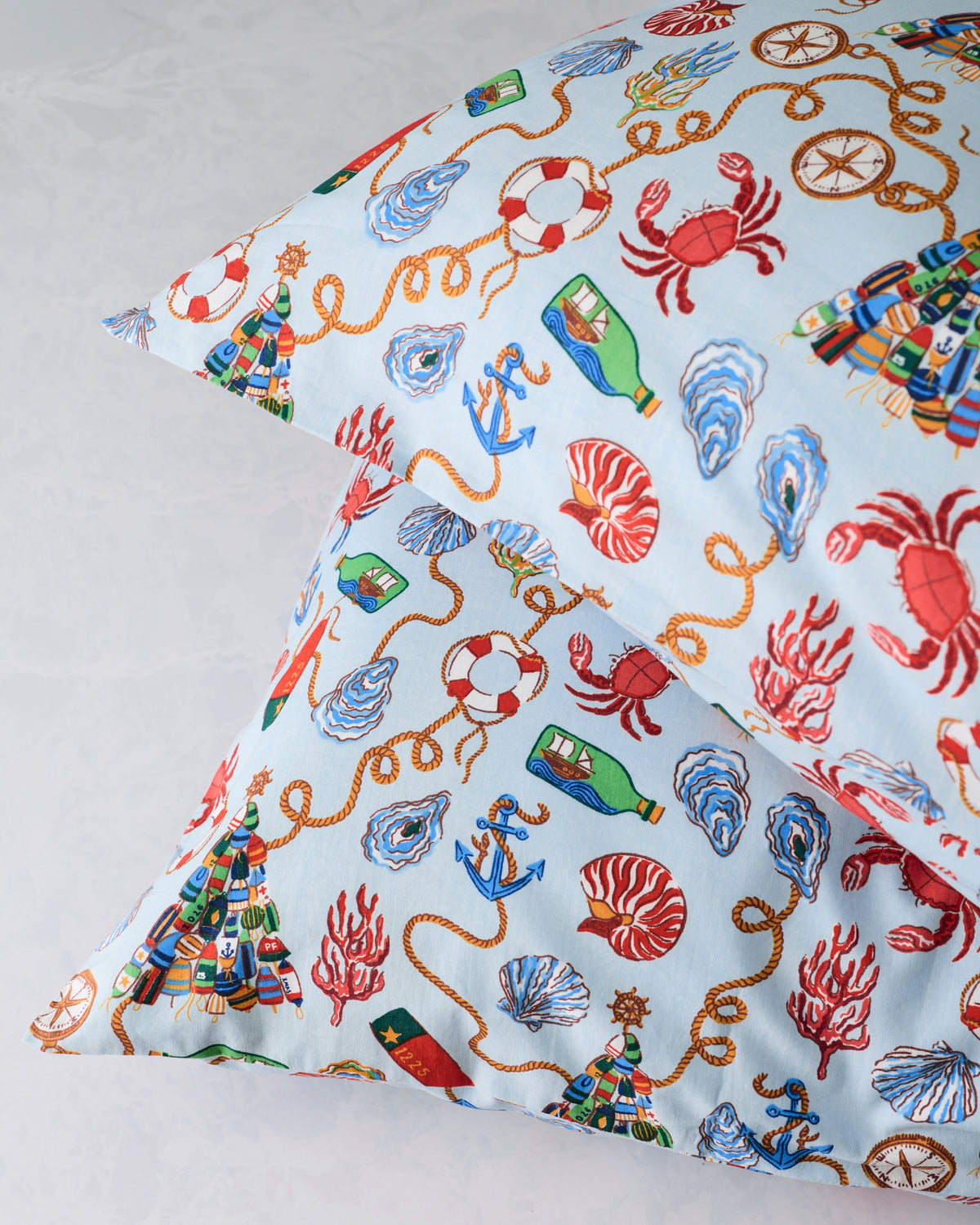 Close-up of two pale blue pillowcases with nautical holiday illustrations, including crabs, shells, anchors, ropes, and lifebuoys.
