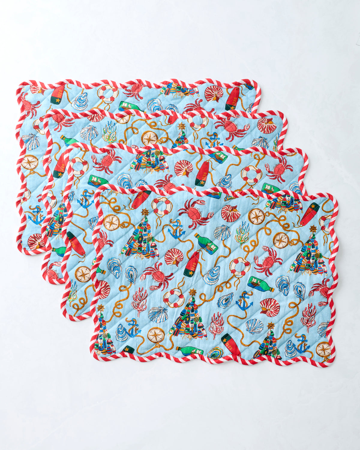 Four blue printed placemats stacked to show their repeating nautical Christmas design and red-and-white scalloped trim.