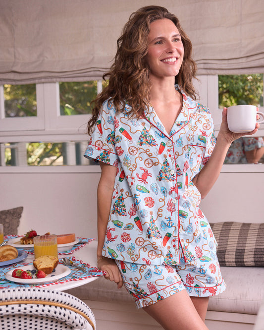Person wearing a light blue nautical-print short pajama set and holding a white cup indoors.