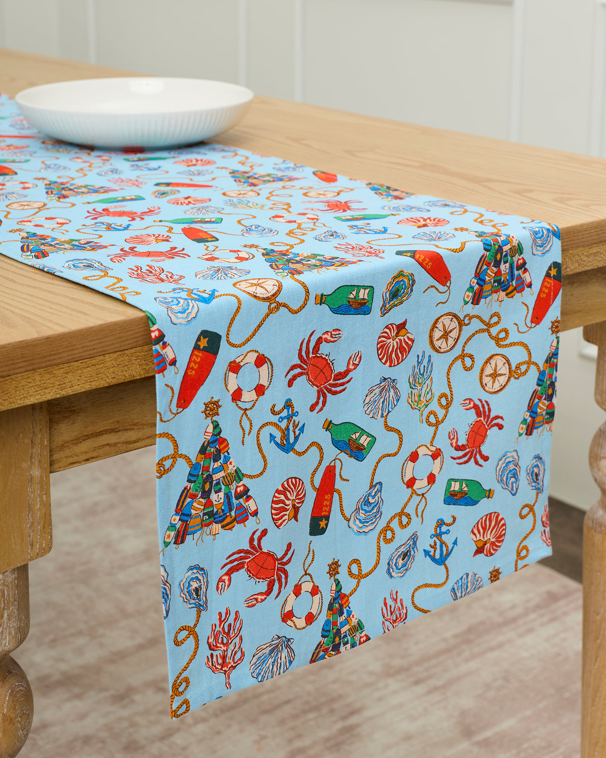 Blue Holidays Ahoy table runner draped over a wooden table, printed with nautical objects, shells, crabs, ropes, and ornaments.
