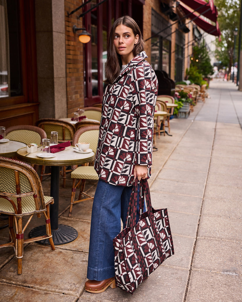 Person standing outside a café wearing a burgundy, cream, and black geometric-patterned jacket and holding a matching tote bag.