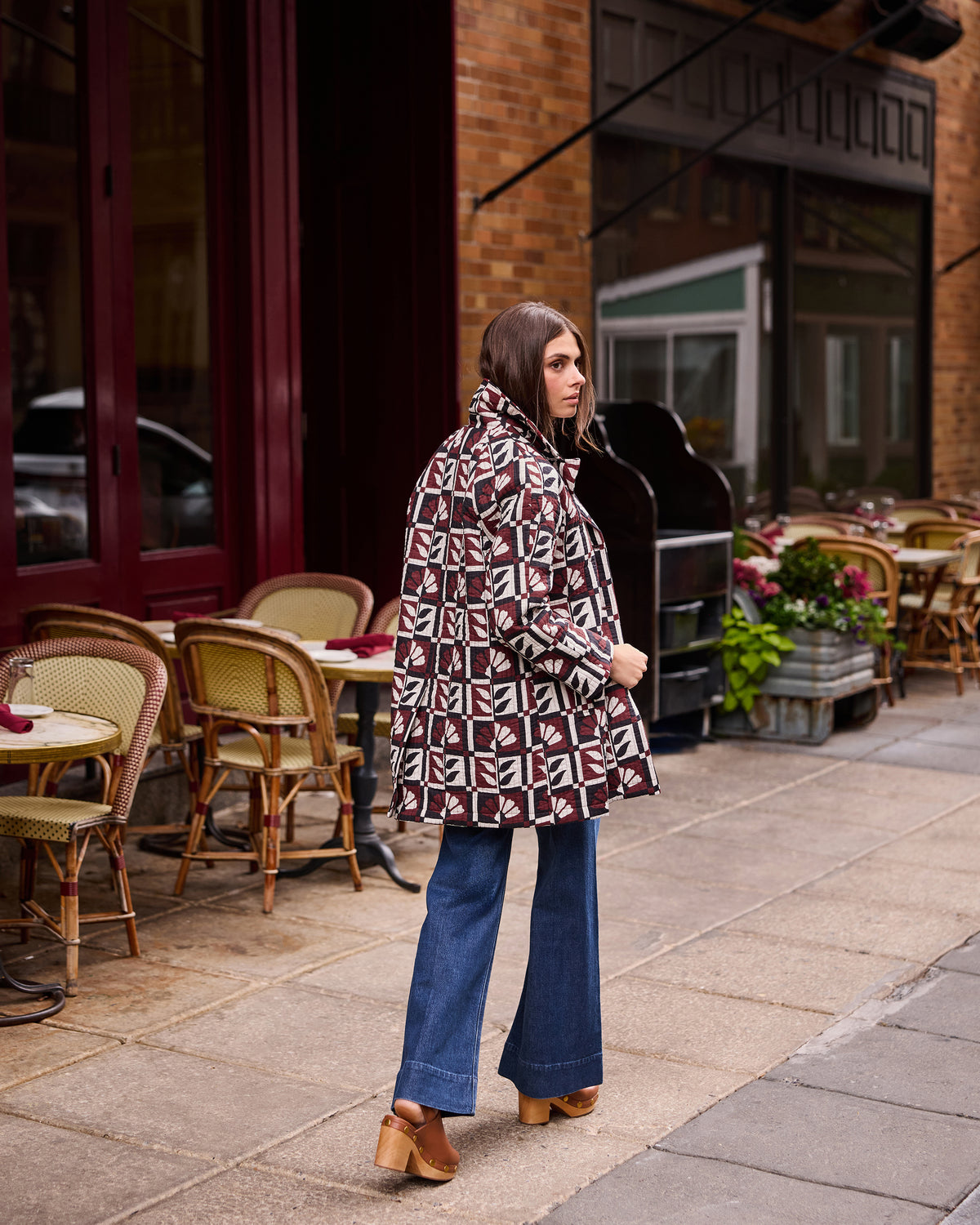 Person shown from behind walking outside a café while wearing a burgundy, cream, and black geometric-patterned Spencer jacket with blue wide-leg jeans.