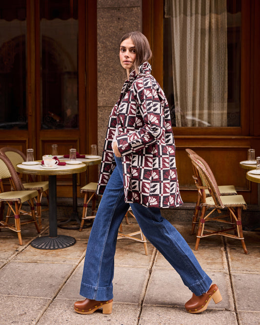 Person walking outside a café wearing a burgundy, cream, and black geometric-patterned Spencer jacket with blue wide-leg jeans.