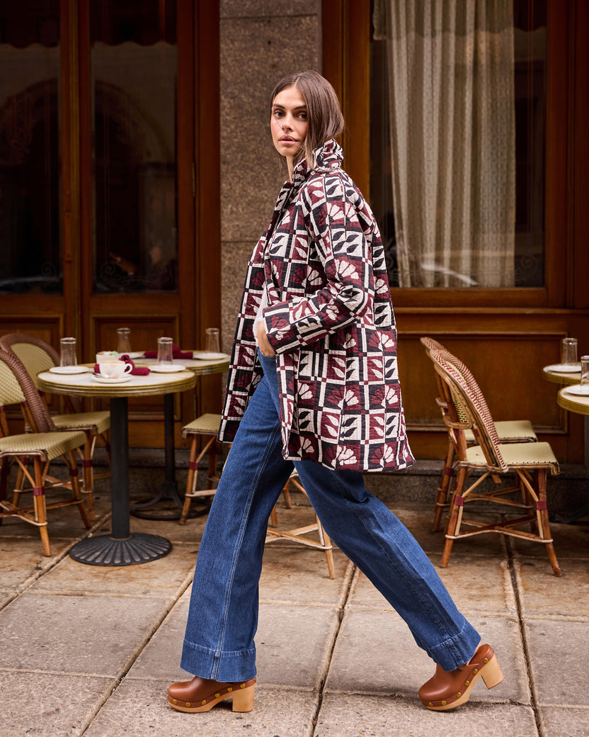 Person walking outside a café wearing a burgundy, cream, and black geometric-patterned Spencer jacket with blue wide-leg jeans.