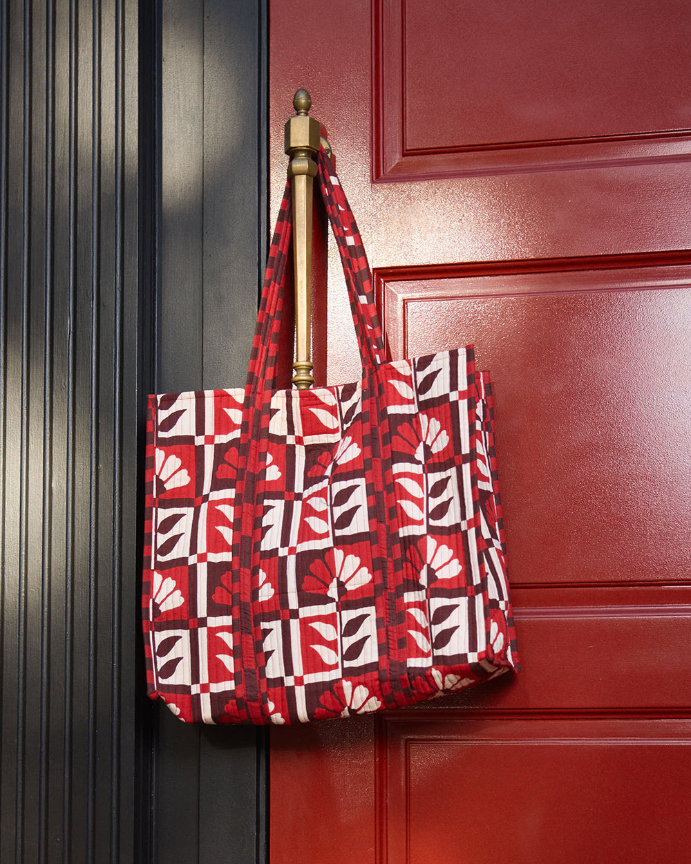 The Traveler Tote hanging from a door handle, featuring a red-and-white floral geometric print and long handles.