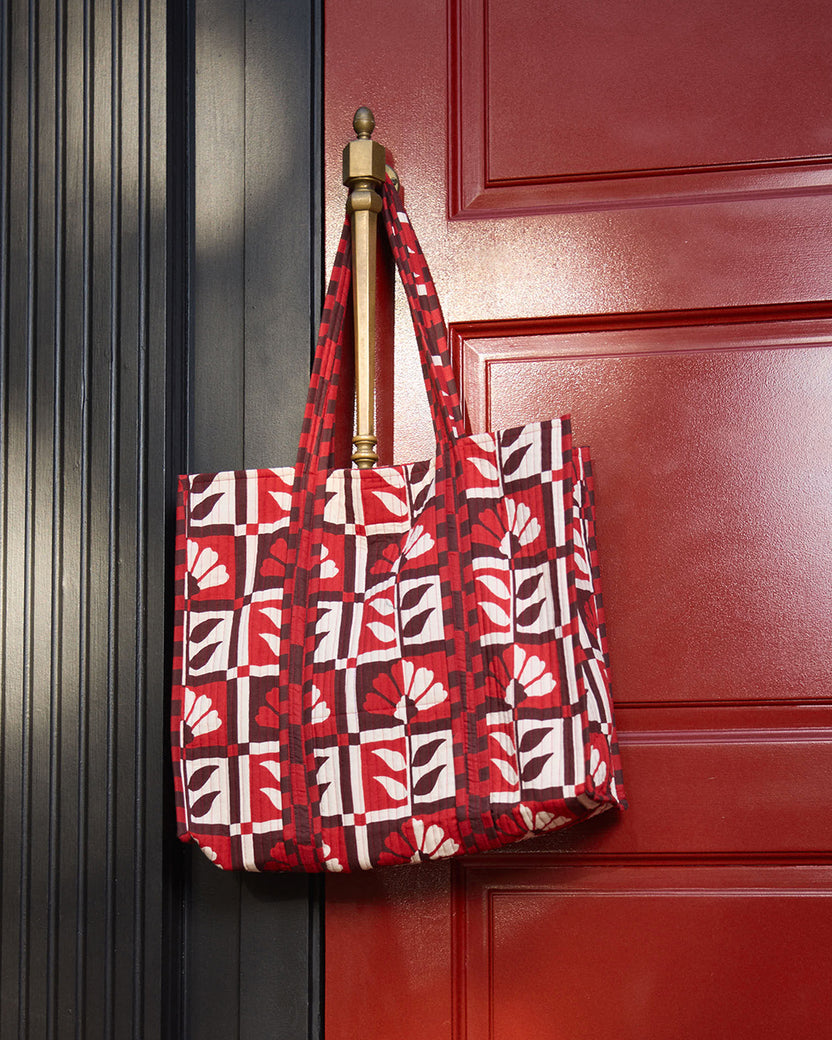 The Traveler Tote hanging from a door handle, featuring a red-and-white floral geometric print and long handles.