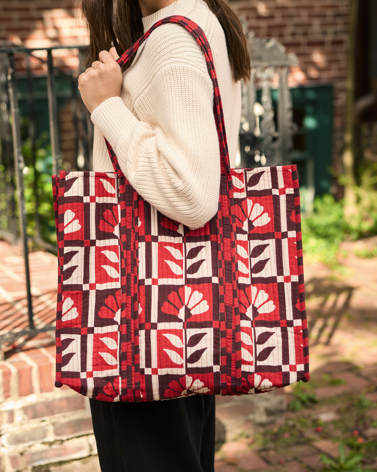The Traveler Tote worn over the shoulder with a cream sweater and black trousers, showing its red-and-white floral geometric print.