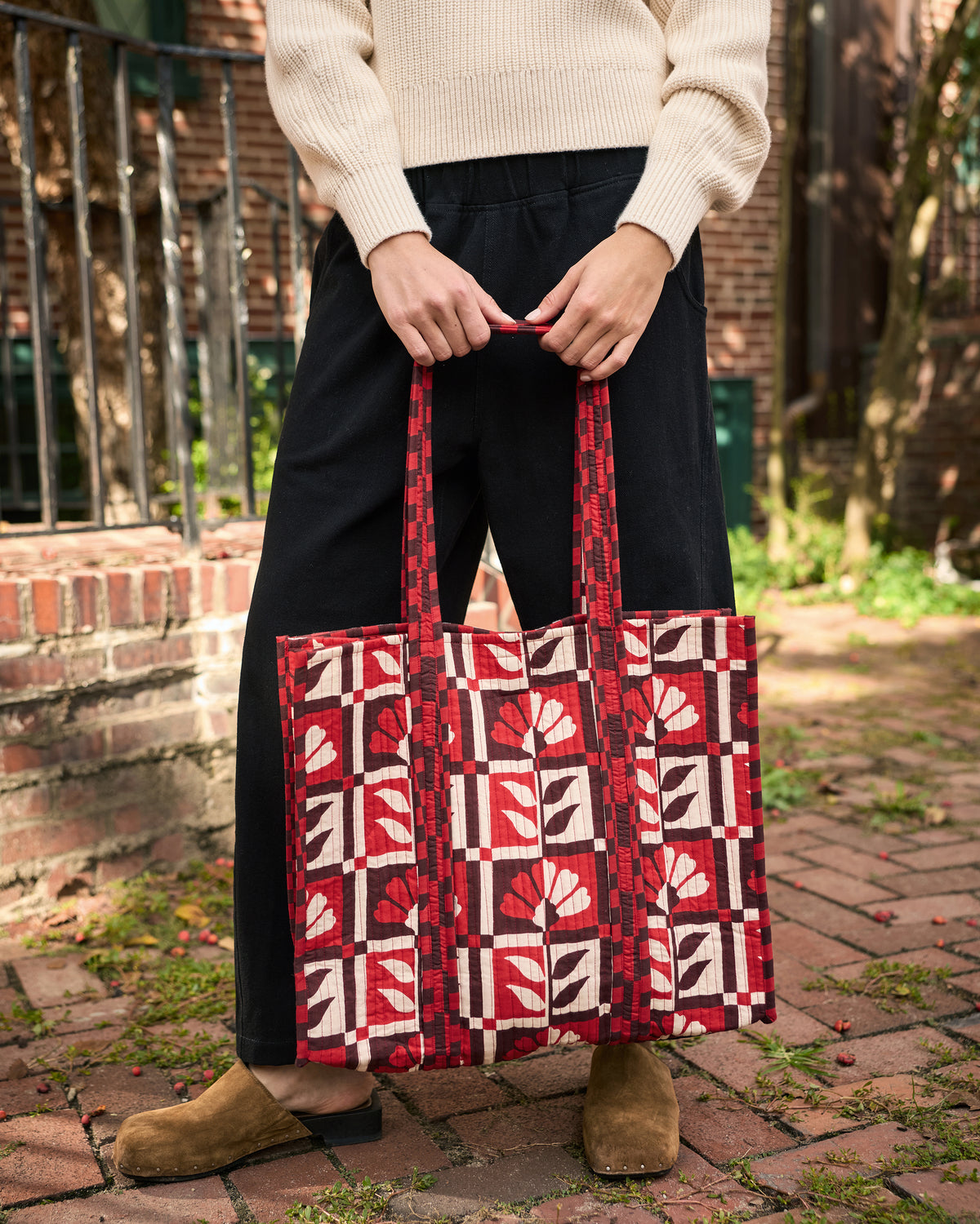 The Traveler Tote held in front of black trousers, showing its red-and-white floral geometric print and long handles.