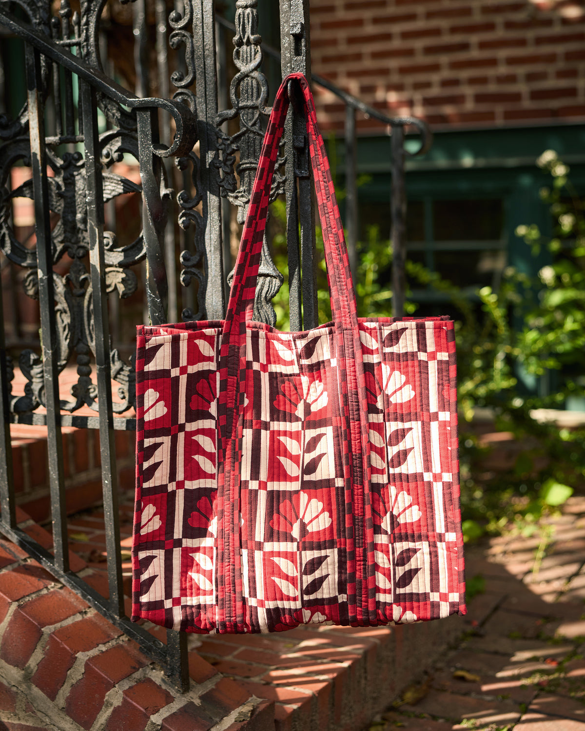 The Traveler Tote hanging from an iron railing, featuring a red-and-white floral geometric print and long handles.