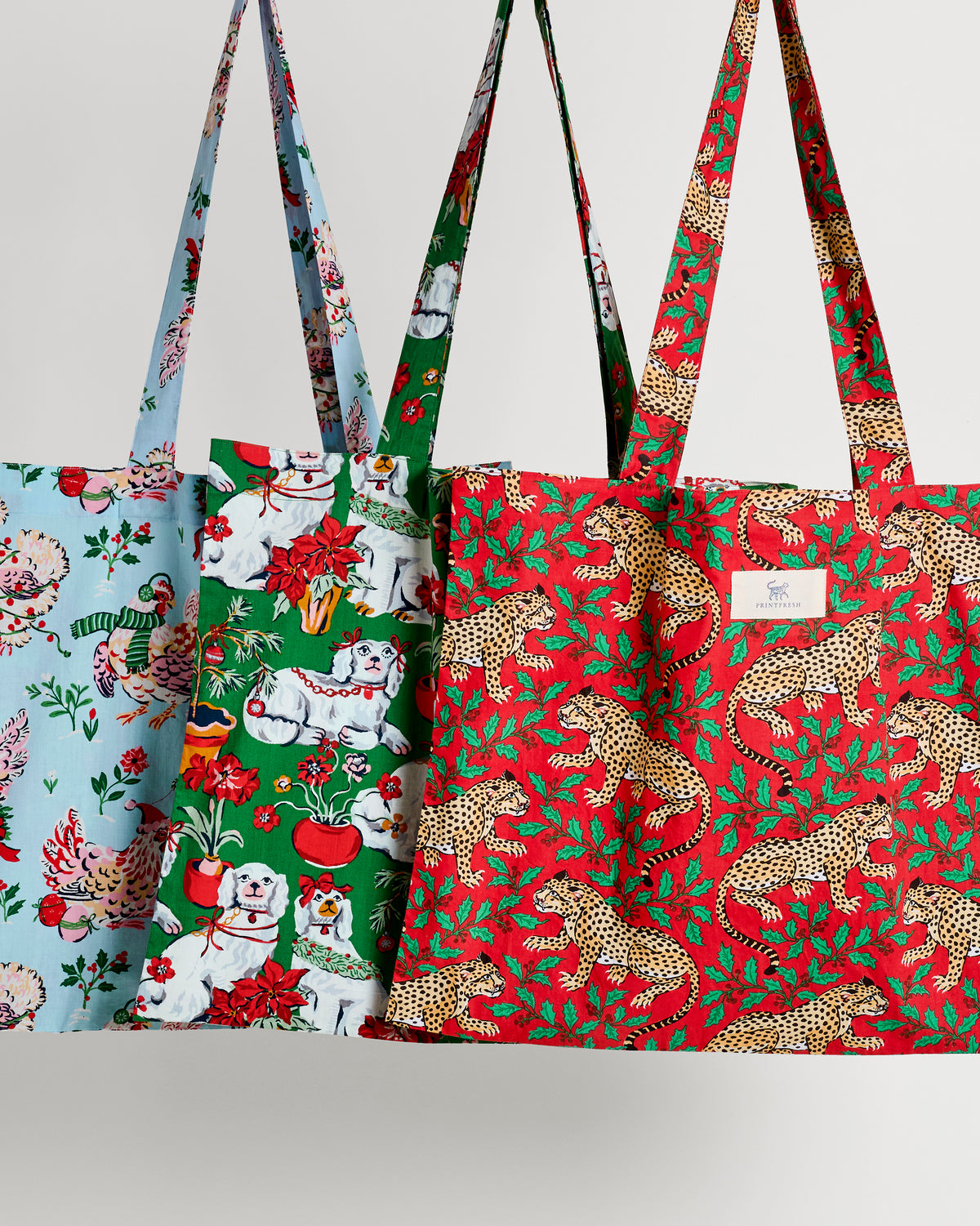 Three patterned tote bags displayed together: pale blue holiday animals, green holiday dogs, and red leopard with holly.