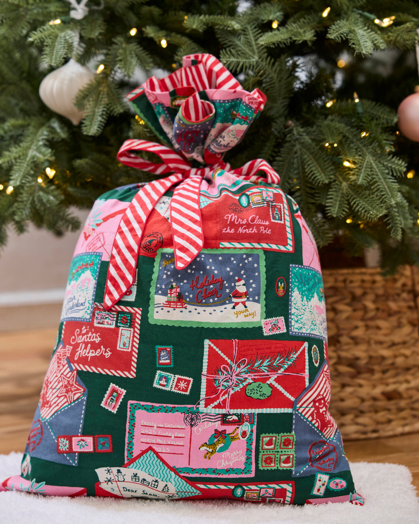 Christmas gift sack tied with a red-and-white striped fabric bow, decorated with illustrated holiday envelopes and postage stamps in front of a lit tree.