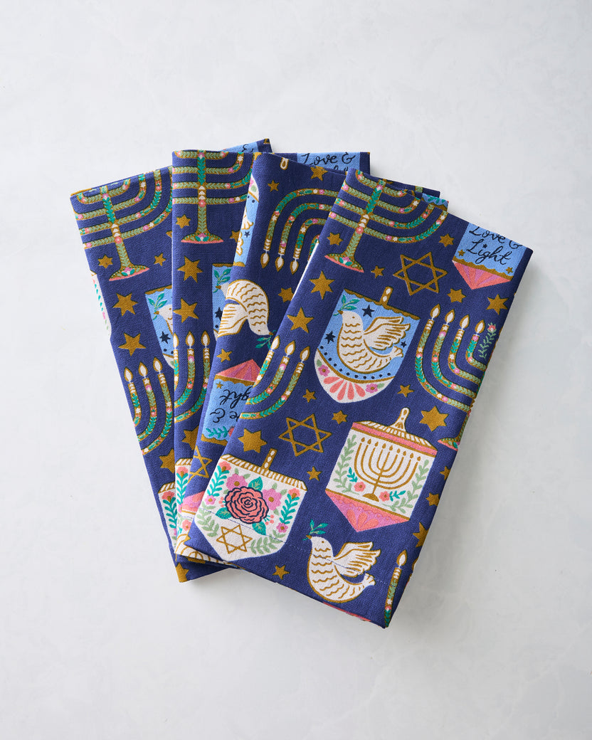 Four folded Hanukkah blue dinner napkins overlapping to show their menorah, dreidel, star, and dove print.