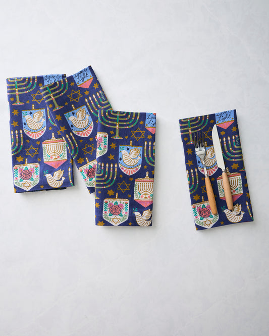 Four folded Hanukkah blue dinner napkins with menorahs, dreidels, stars, and dove motifs; one napkin holds a fork and knife.