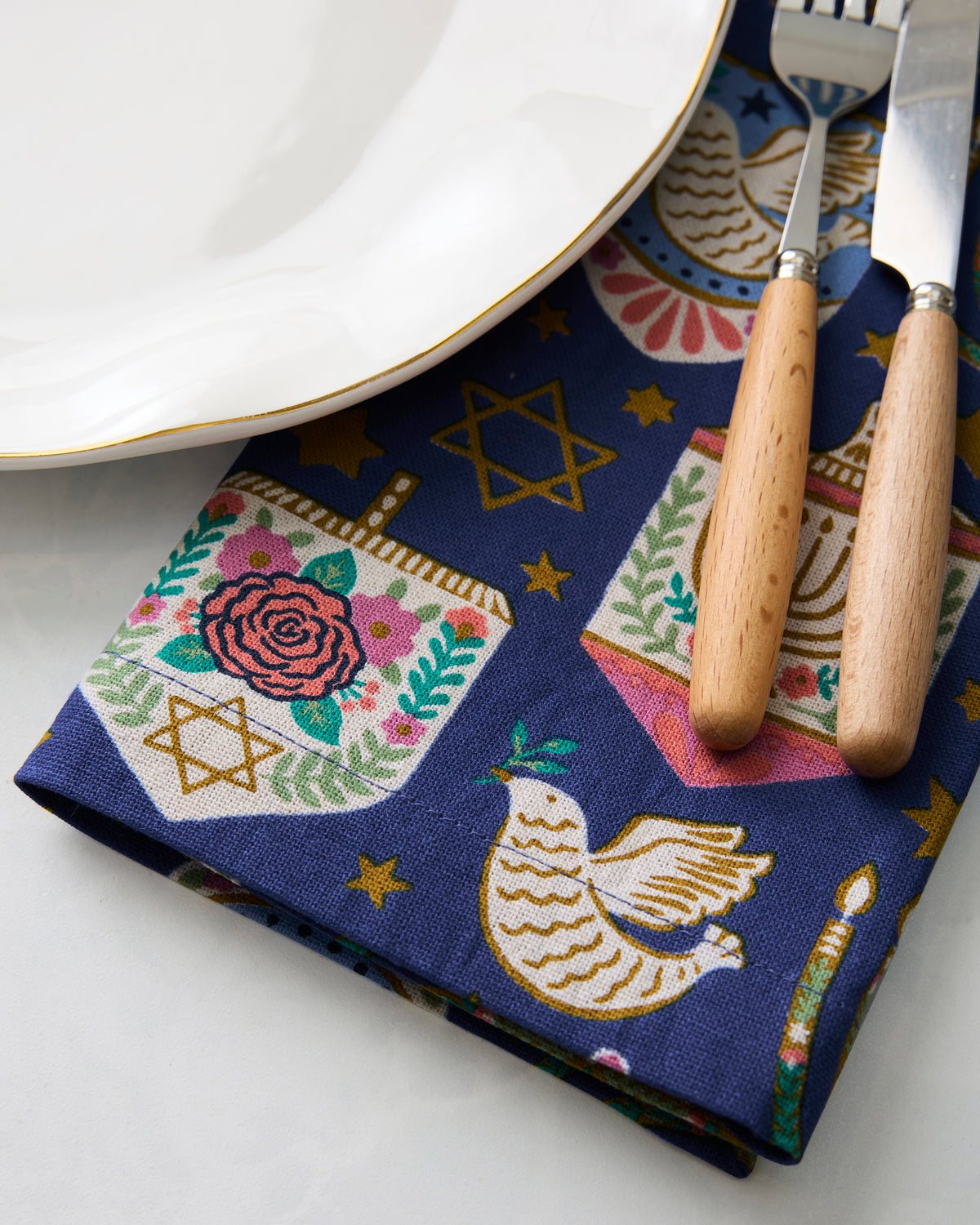Close-up of a Hanukkah blue dinner napkin beneath a white plate, fork, and knife, showing the menorah, dove, star, and floral motif.