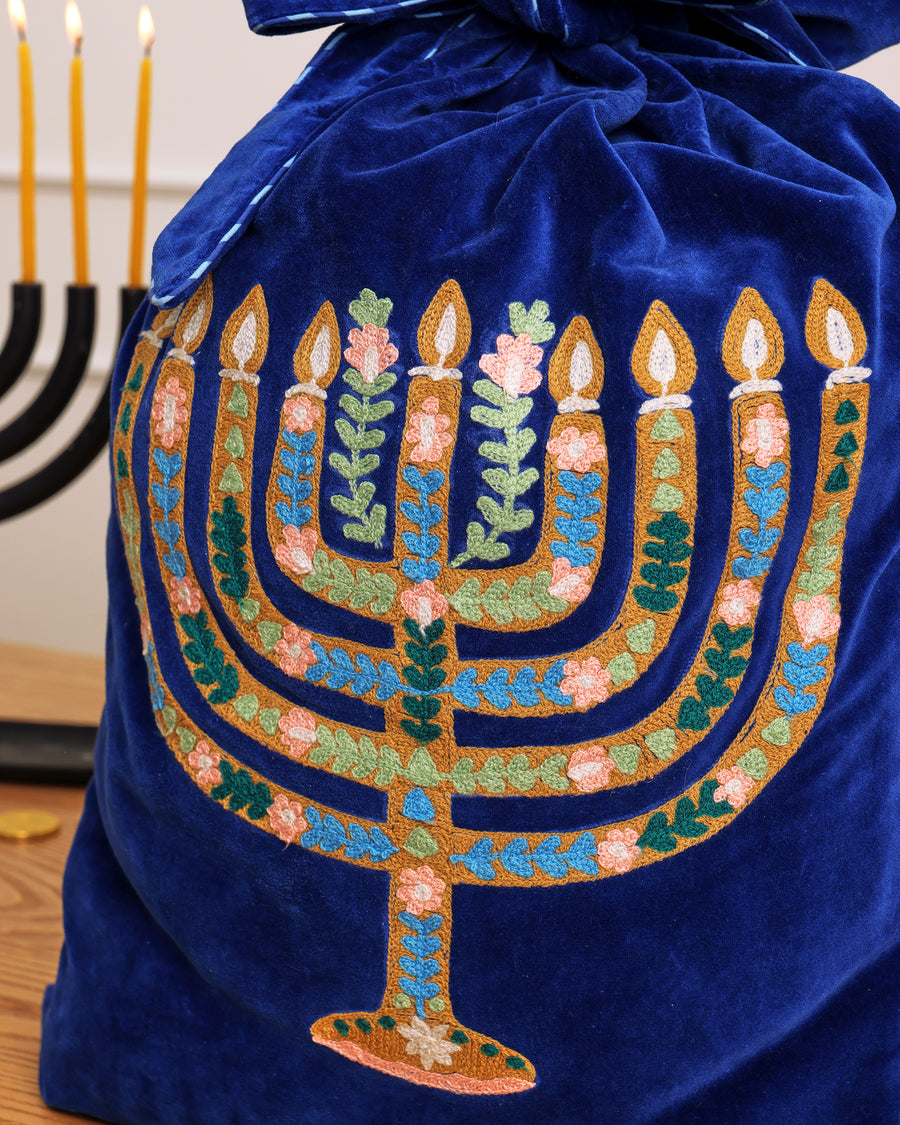 Blue gift sack with colorful embroidered Hanukkah menorah and floral motifs, shown beside a lit menorah.