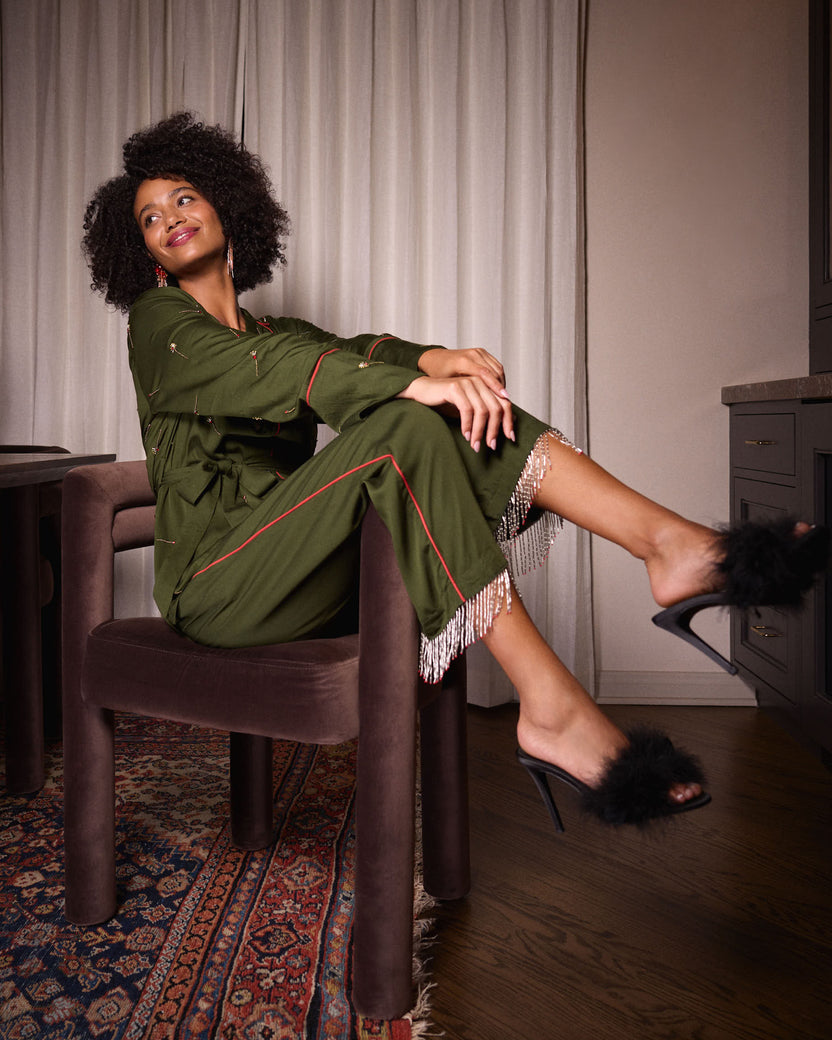 Seated view of the olive Matchmaker pajama set showing the belted top, piped pants, beaded tassel hems, and black feather-trimmed heeled shoes.