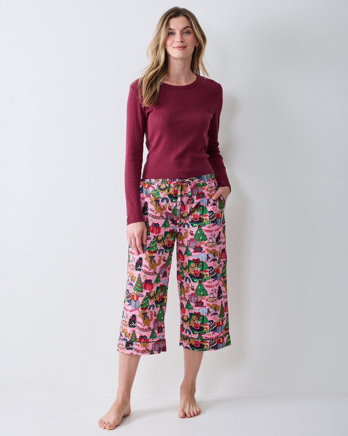Person wearing candy pink cropped pajama pants with a colorful Christmas cat-and-gifts print, shown with a burgundy ribbed top.