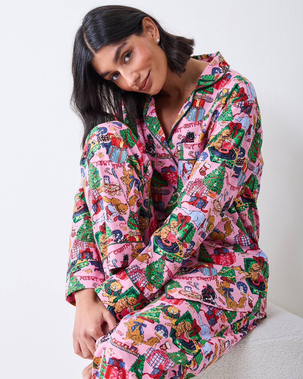 Seated model wearing a candy-pink long pajama set with a festive cat-and-gift print.
