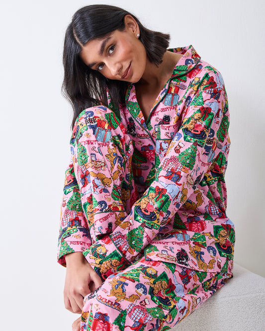 Seated model wearing a candy-pink long pajama set with a festive cat-and-gift print.