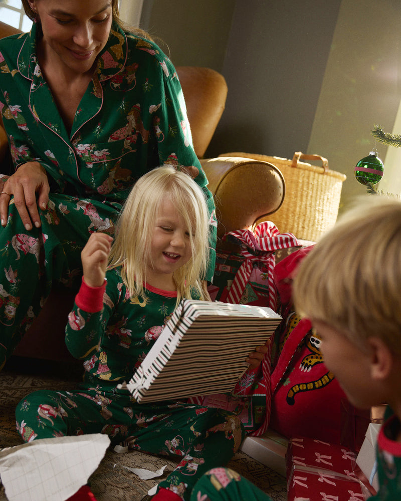Two people and two children sit together indoors near a decorated Christmas tree, wearing green and red printed pajamas.