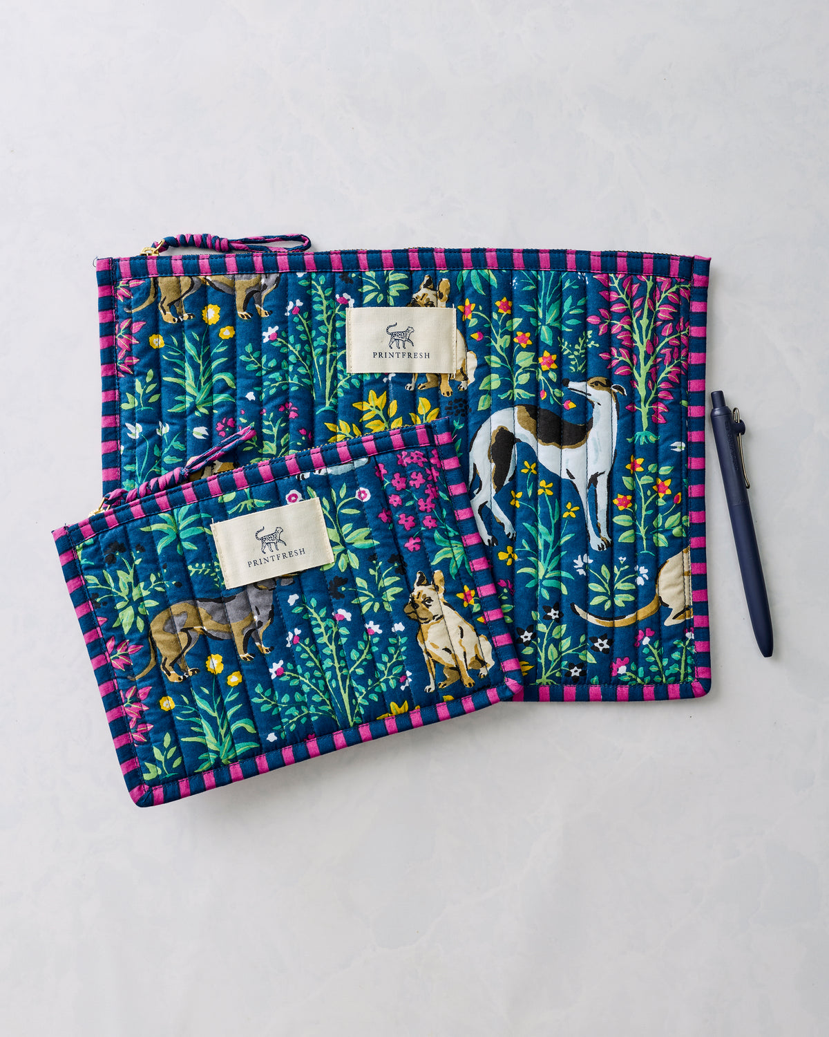 Two indigo Must Love Dogs flat pouches with pink striped trim, shown beside a pen.