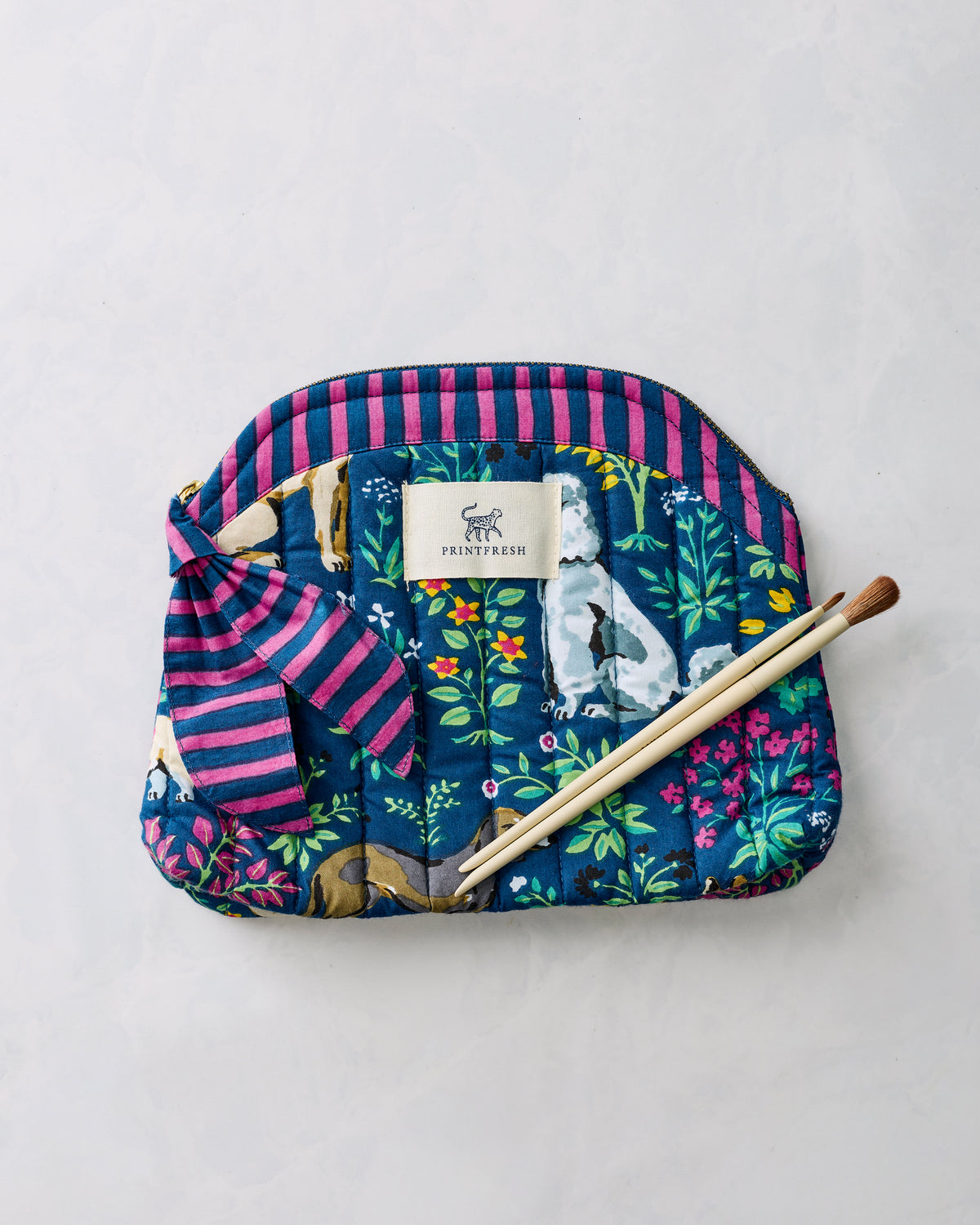Indigo quilted makeup bag with pink-and-blue striped trim, colorful floral and dog print, Printfresh label, and two makeup brushes placed across it.
