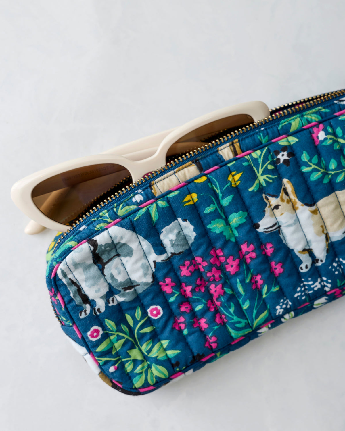 Quilted indigo zippered sunglasses case with floral and dog print, shown with sunglasses partially inserted.
