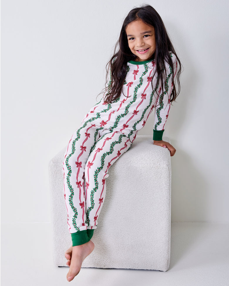 Child wearing the Ribbons & Bows Vanilla Cream kids long pajama set while seated on a white upholstered block.