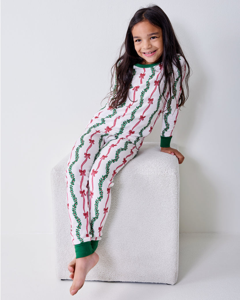 Child wearing the Ribbons & Bows Vanilla Cream kids long pajama set while seated on a white upholstered block.
