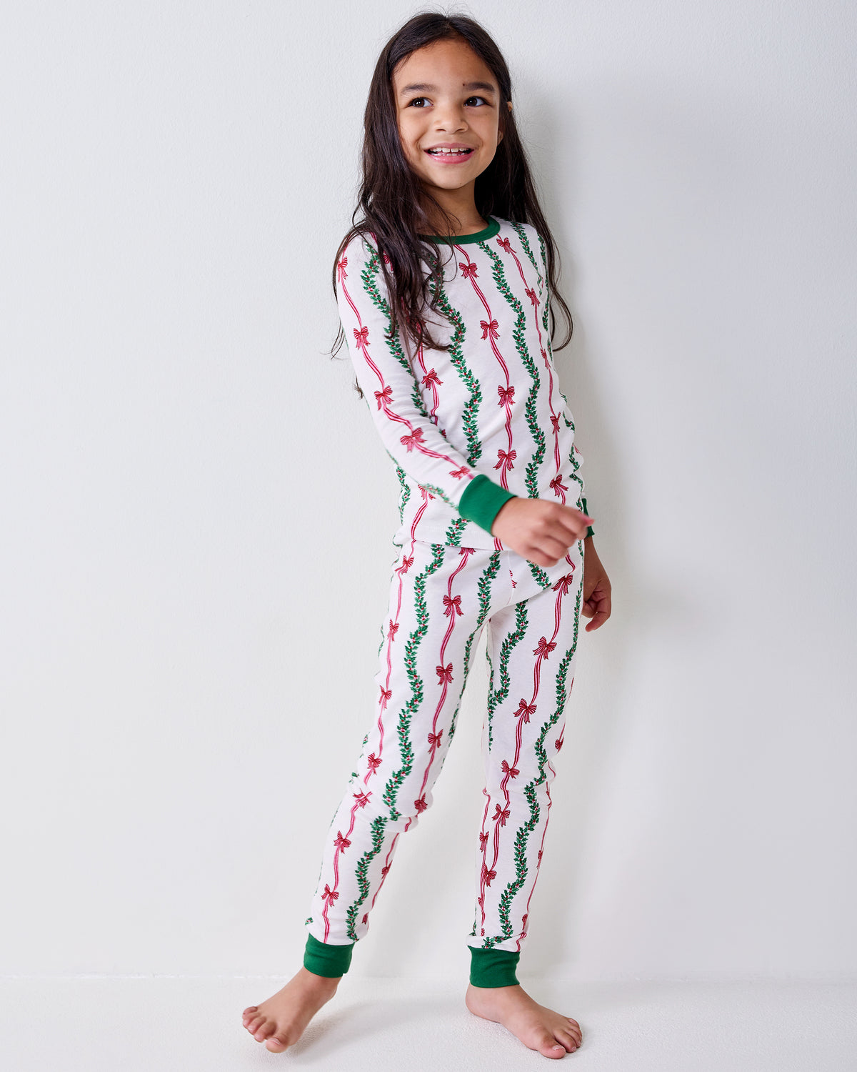 Child wearing the long-sleeve Ribbons & Bows pajama set with green cuffs and pink bow and garland print.