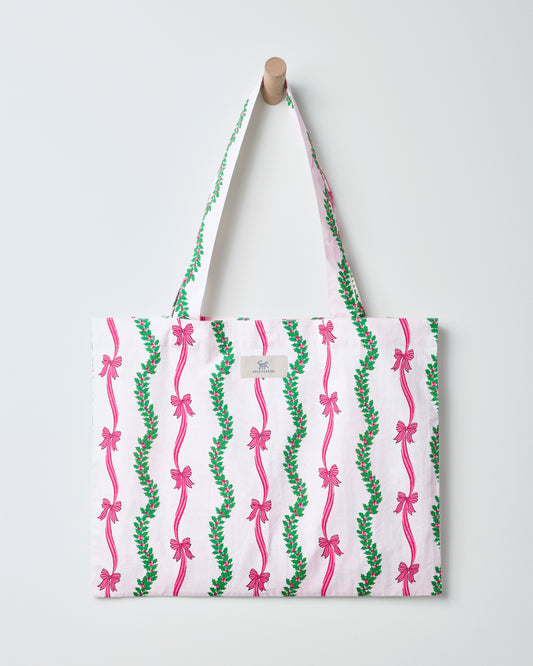 Ribbons & Bows cotton tote bag hanging from a wall peg, with a white background and vertical green garland and pink bow pattern.