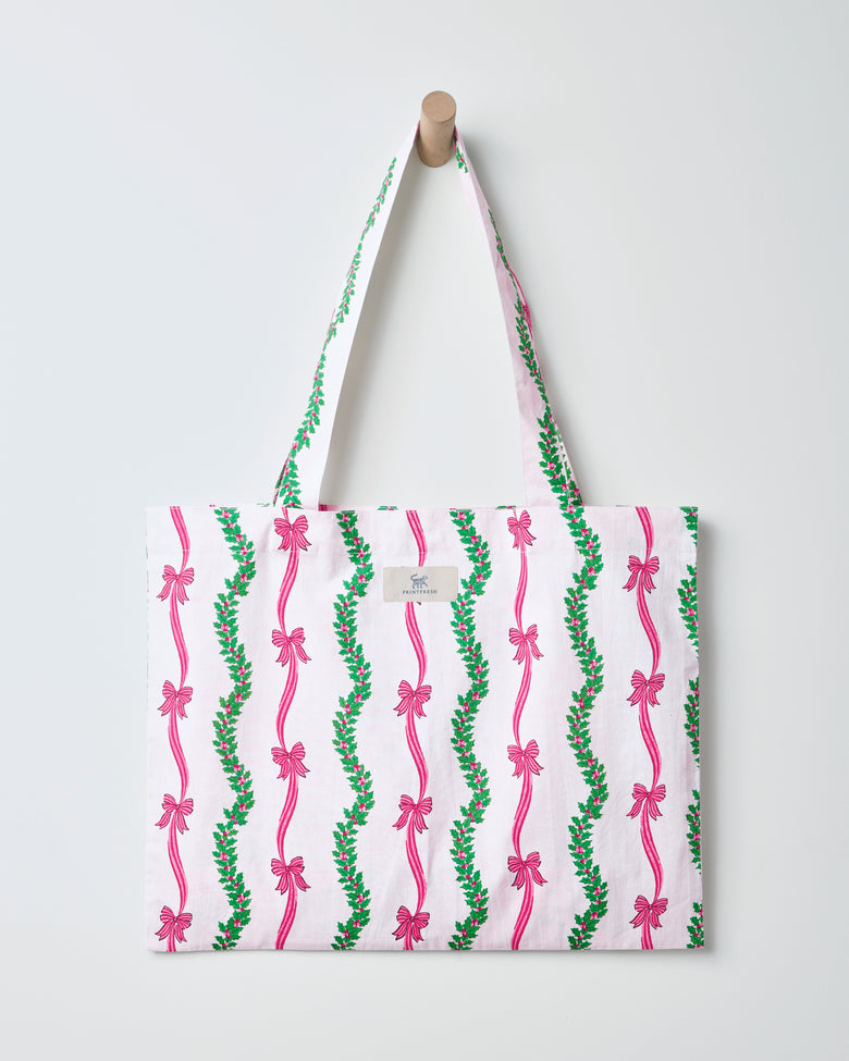 Ribbons & Bows cotton tote bag hanging from a wall peg, with a white background and vertical green garland and pink bow pattern.