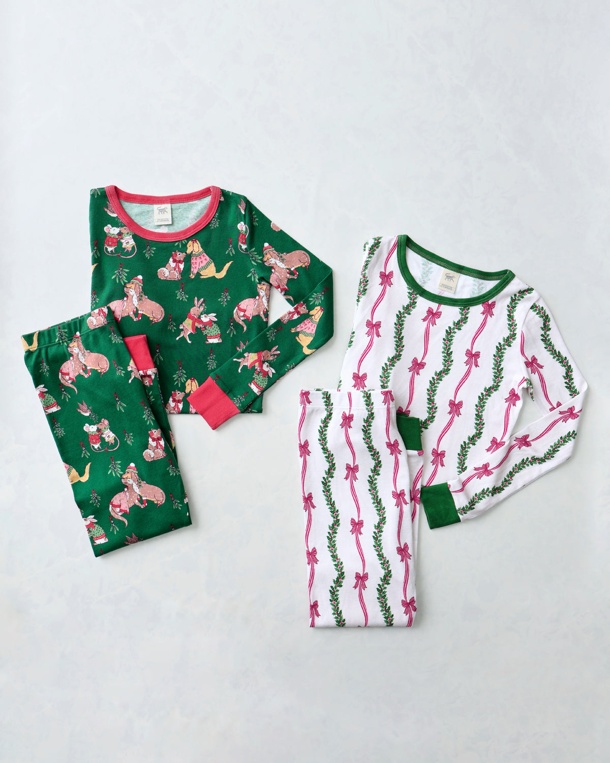 Flat lay of two children's long-sleeve pajama sets: a green festive animal-print set and a white ribbon-and-mistletoe set.