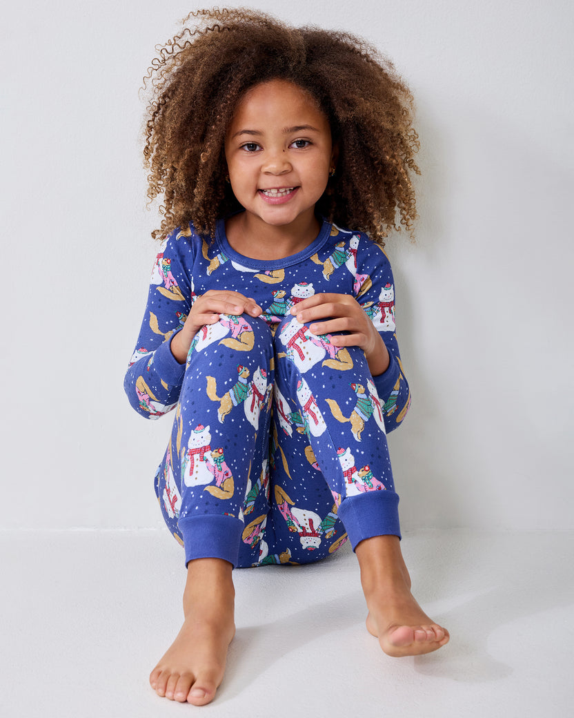 Child seated on the floor wearing blue long-sleeve pajamas with a snowman, cat, and winter print.