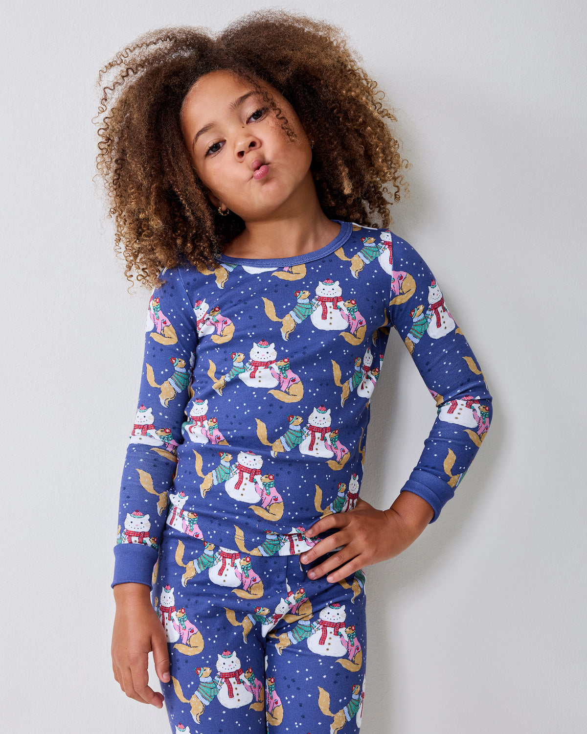 Child standing against a wall wearing blue long-sleeve pajamas with a snowman, cat, and winter print.