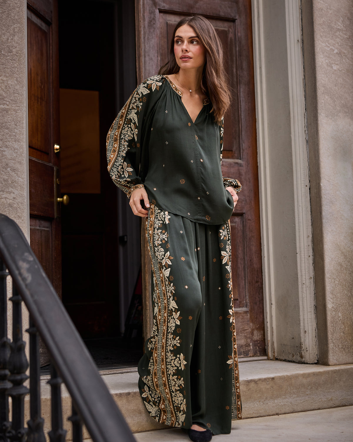 Full-length front view of the Hallie Blouse and Sasha Pants in dark green with floral patterned side panels, standing in a doorway.