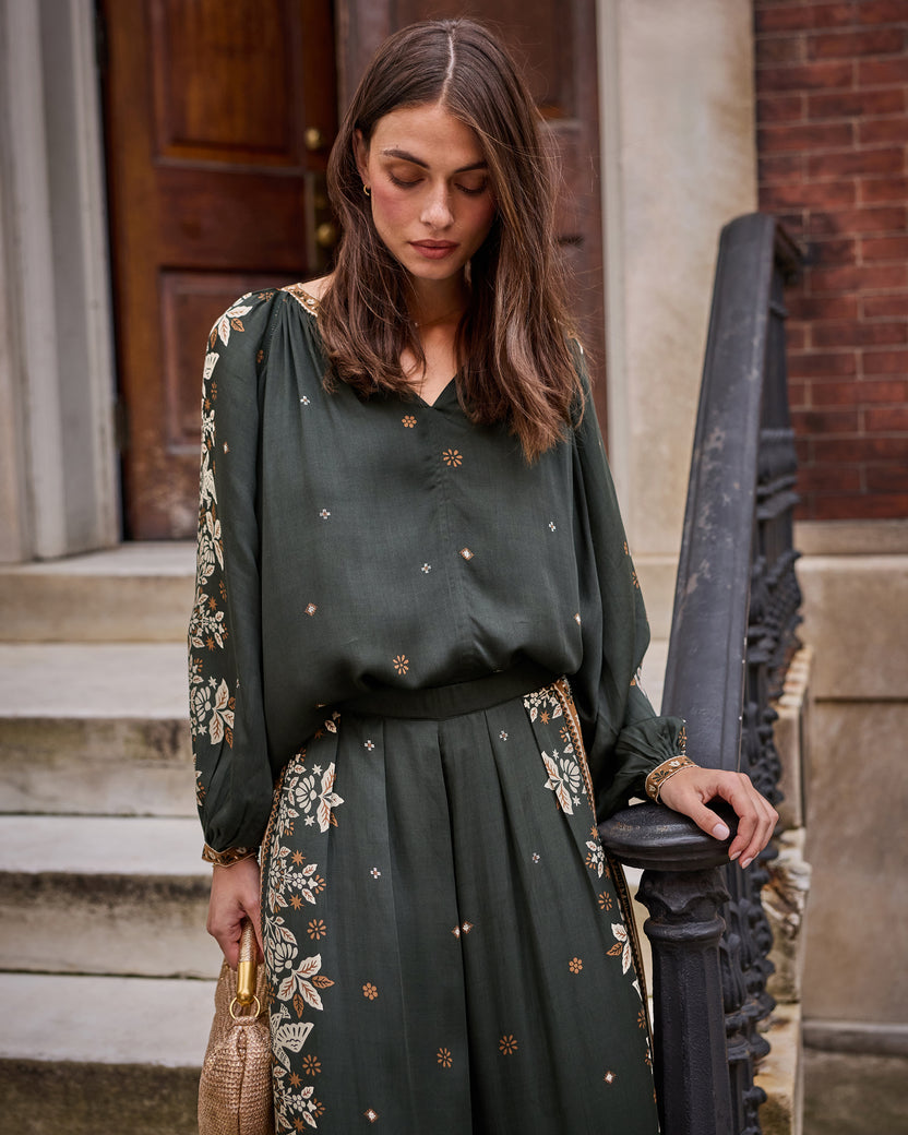 Front view of the Hallie Blouse in dark green with a V-neck, scattered motifs, and floral patterned sleeves, worn on outdoor steps.