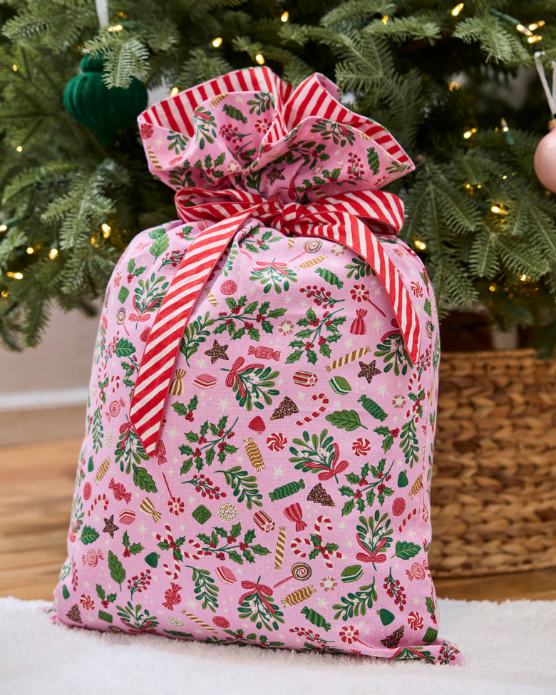 Pink patterned gift sack tied with a large red-and-white striped bow in front of a Christmas tree.
