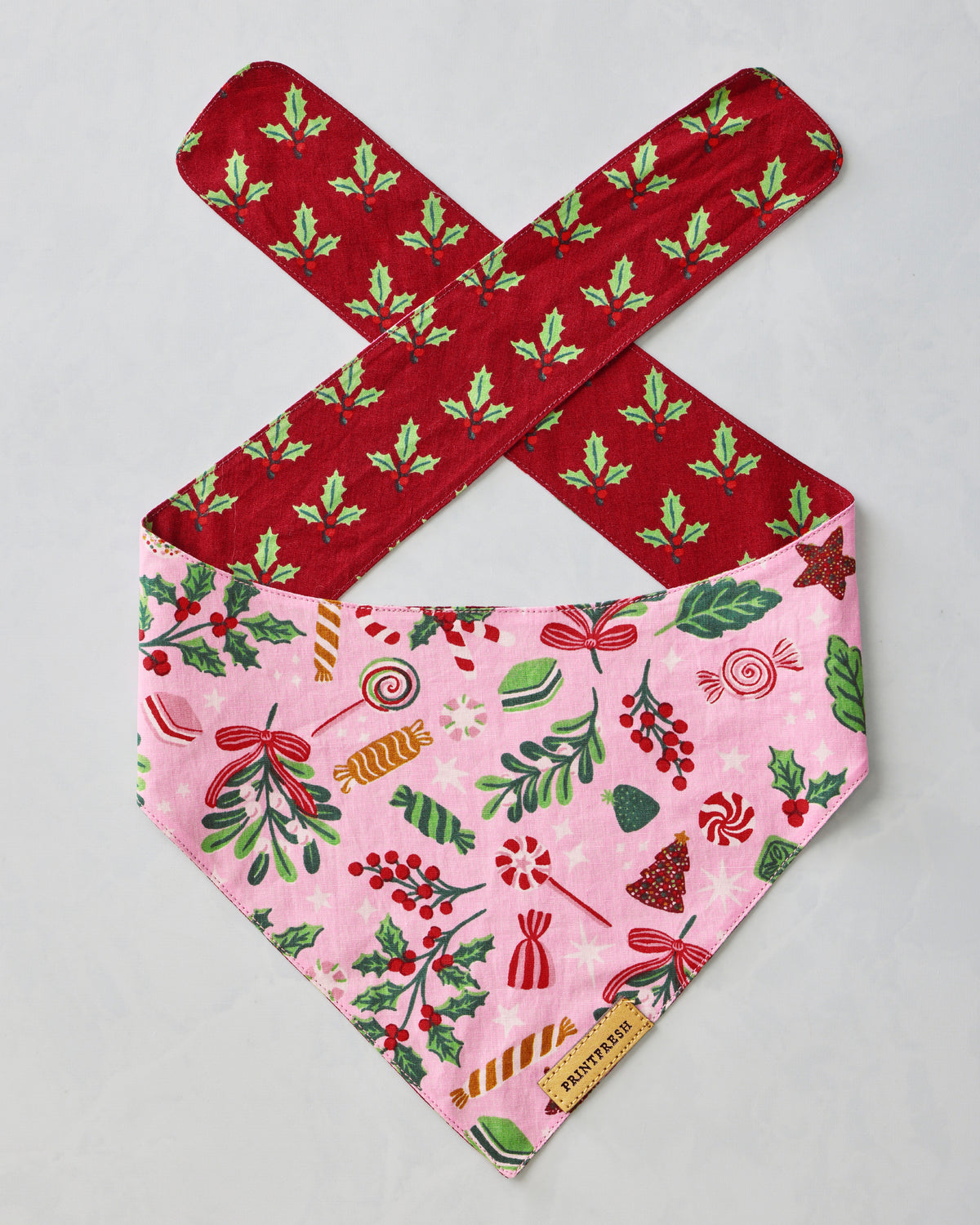 Pet bandana laid flat with a pink candy-patterned front and red patterned ties; a brand label is attached at the point.