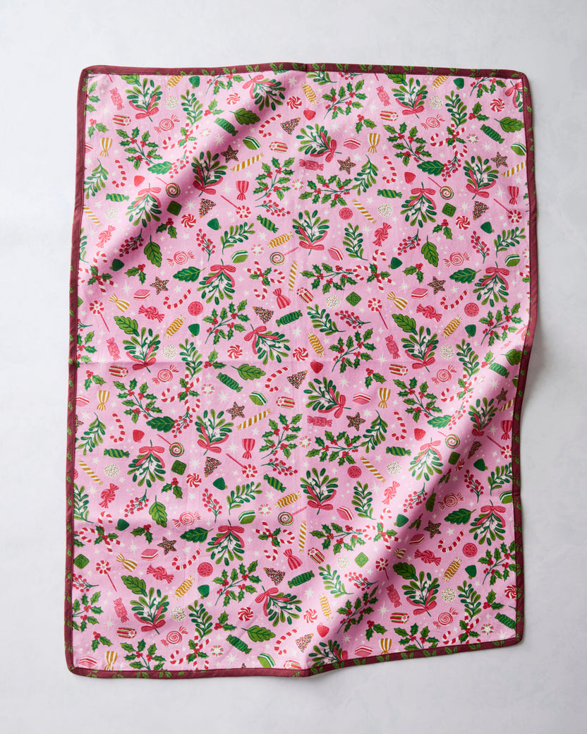 Tea towel with a pink holiday candy, holly, and foliage print, shown flat with a folded corner.