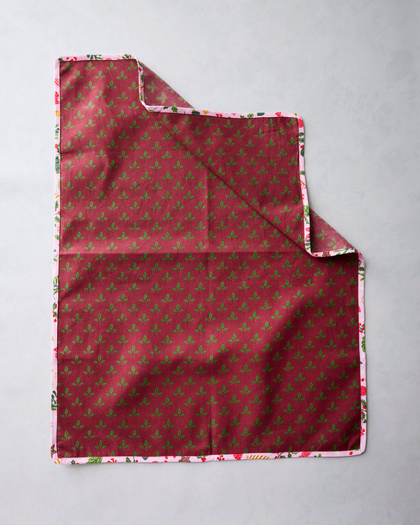 Tea towel showing the burgundy reverse side with a repeating green sprig pattern and the printed edge visible around it.