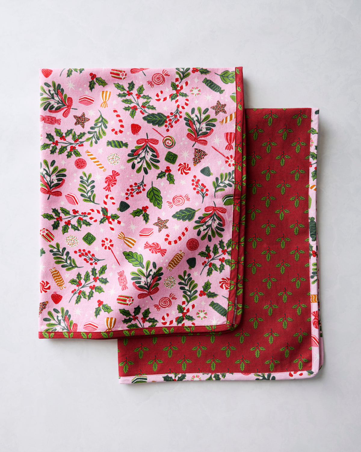 Two tea towels displayed together, showing the pink holiday print on one towel and the burgundy sprig-patterned reverse on the other.