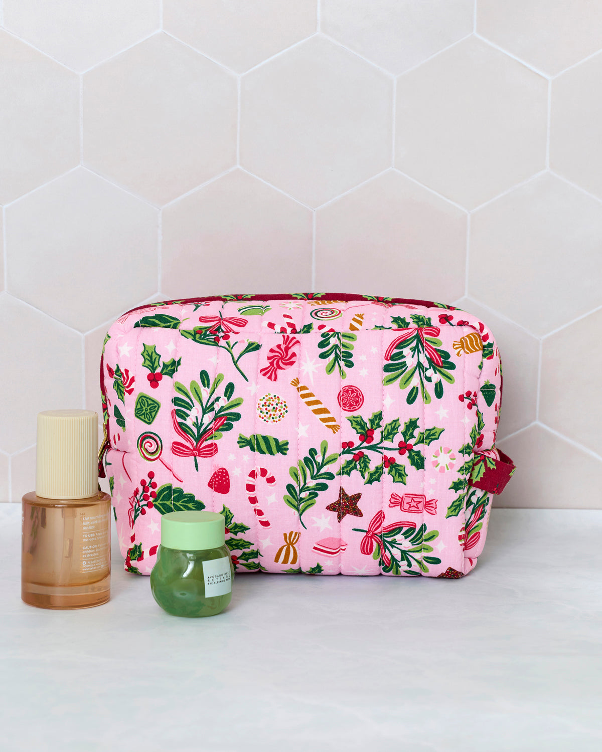 Candy pink quilted pouch with a colorful holiday print, shown upright beside two cosmetic containers for scale.