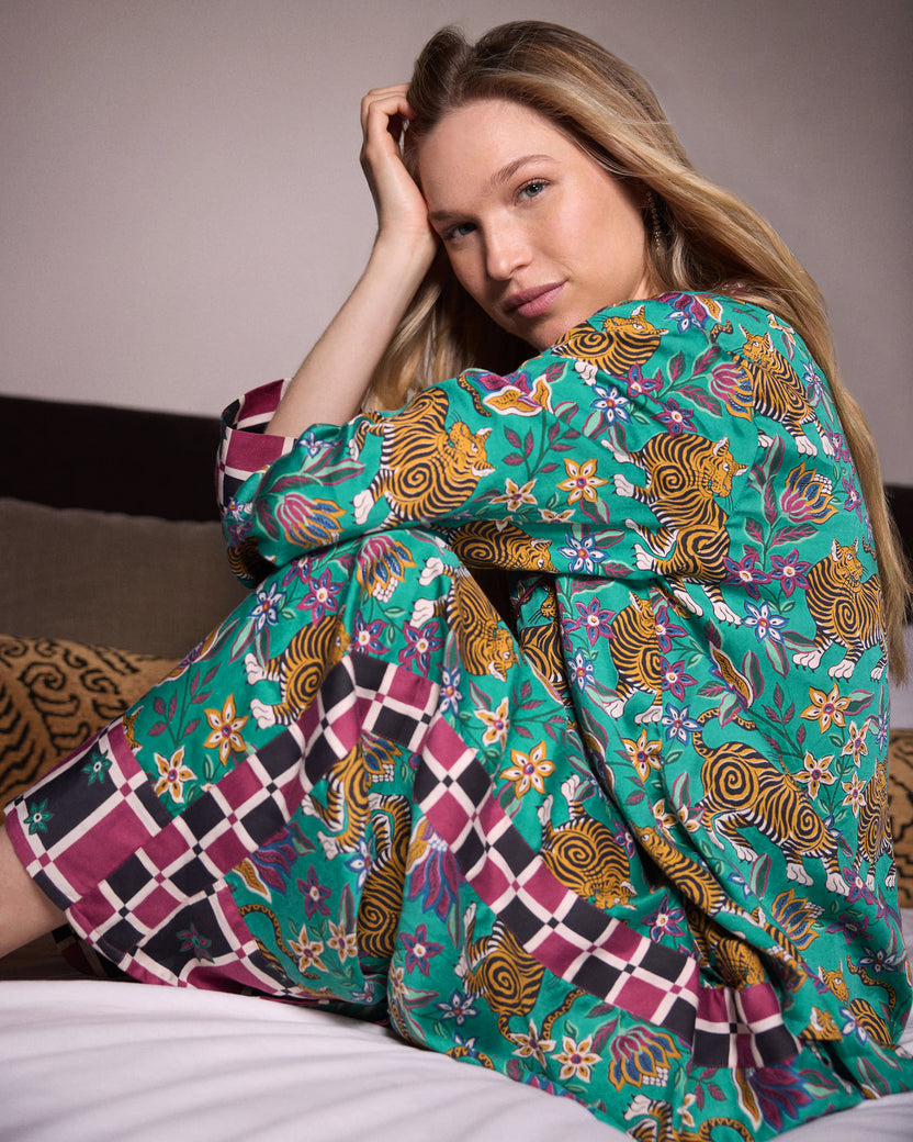 Side view of a person seated on a bed wearing the Tibetan Tigress pajama shirt and pants set with a teal tiger and floral print and checked trim.