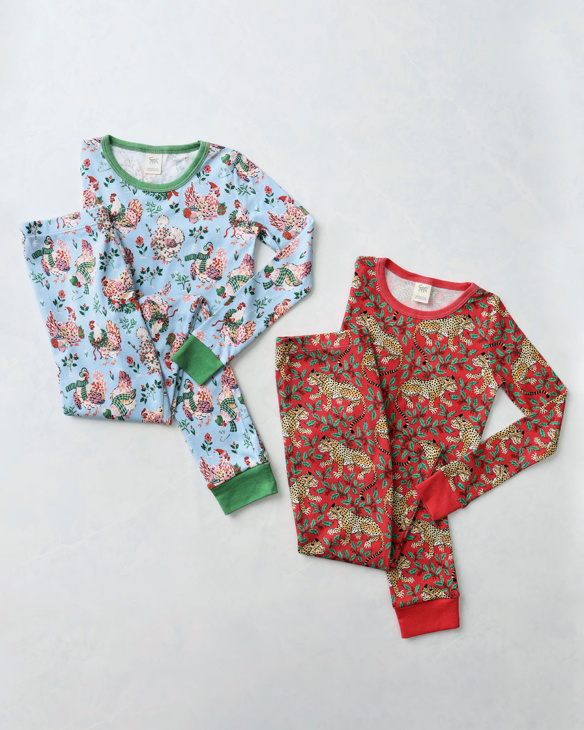 Flat lay of two children's long-sleeve pajama sets: a light blue hen-print set and a red leopard-print set.