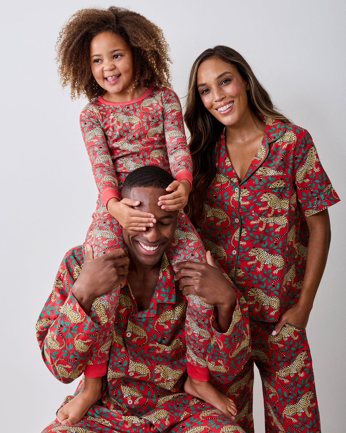 Child wearing a ruby Winter Holly Bagheera long pajama set sits on an adult's shoulders; both wear matching leopard-and-holly prints.
