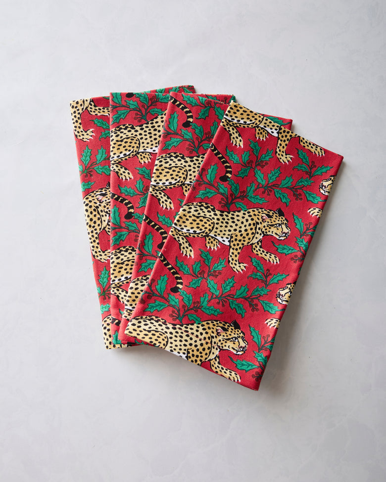 Four folded ruby dinner napkins with leopard and holly print arranged in an overlapping stack.