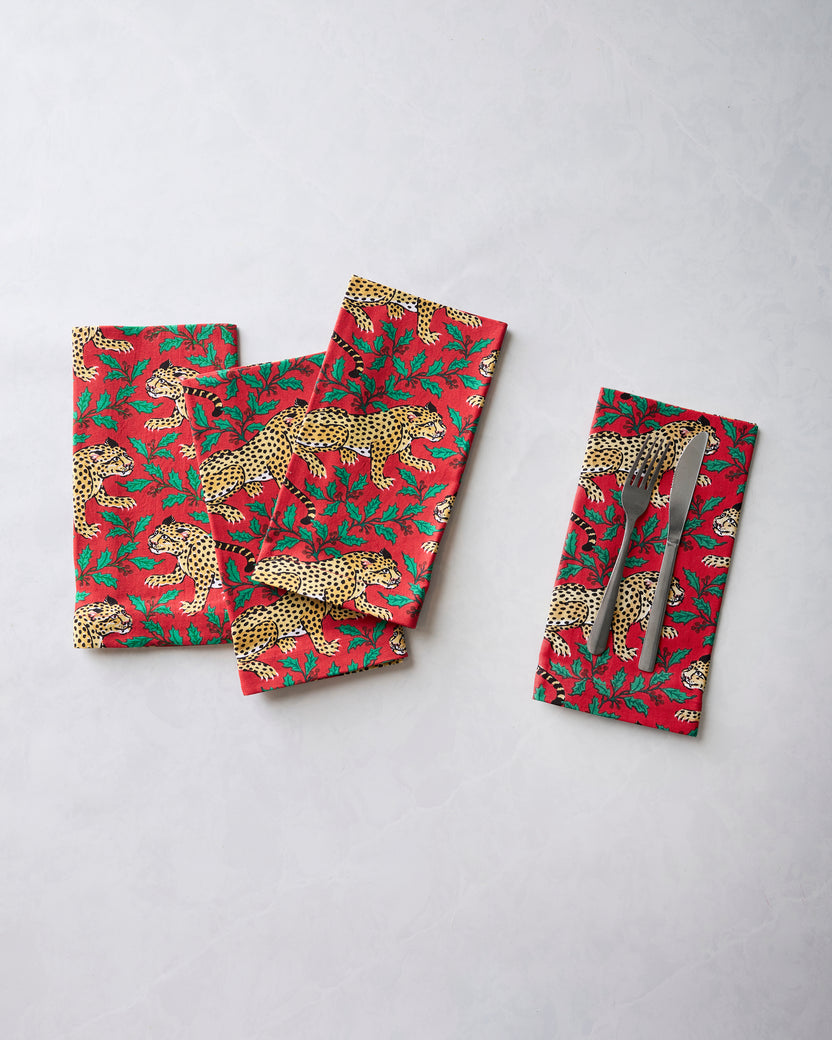 Four ruby leopard-and-holly printed dinner napkins displayed with a separate napkin holding a fork and knife.